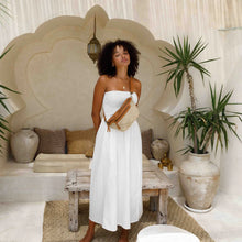 Load image into Gallery viewer, Woman in a white dress standing in a stylish indoor setting with plants and a wooden ceiling.
