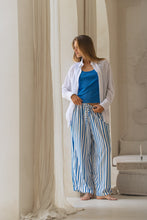 Load image into Gallery viewer, Woman wearing a white shirt, blue tank top, and striped pants standing in a softly lit room.