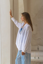 Load image into Gallery viewer, Woman wearing a white shirt, blue top, and striped pants standing in a softly lit room.