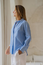 Load image into Gallery viewer, Woman wearing a blue striped shirt and white pants in a room with a lounge in the background