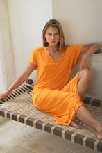 Load image into Gallery viewer, Woman in an orange dress sitting on a ledge with a neutral background