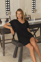 Load image into Gallery viewer, Woman in a black mini dress sitting on a wooden stool in a room with a mirror and decorative items.