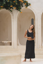 Load image into Gallery viewer, Woman in a black maxi skirt and tank top standing in a marble archway with floral decorations.