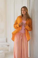Load image into Gallery viewer, Woman wearing an orange shirt over a pink top and orange striped pants leaning against a white wall.