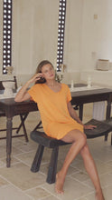 Load and play video in Gallery viewer, Woman in an orange dress sitting on a wooden bench in a room with a table and decorative items.
