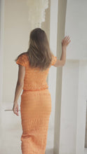 Load and play video in Gallery viewer, Woman in orange shirred top and maxi skirt standing on steps with a decorative wall in the background