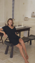 Load and play video in Gallery viewer, Woman in a black mini dress sitting on a wooden stool in a room with a mirror and decorative items.