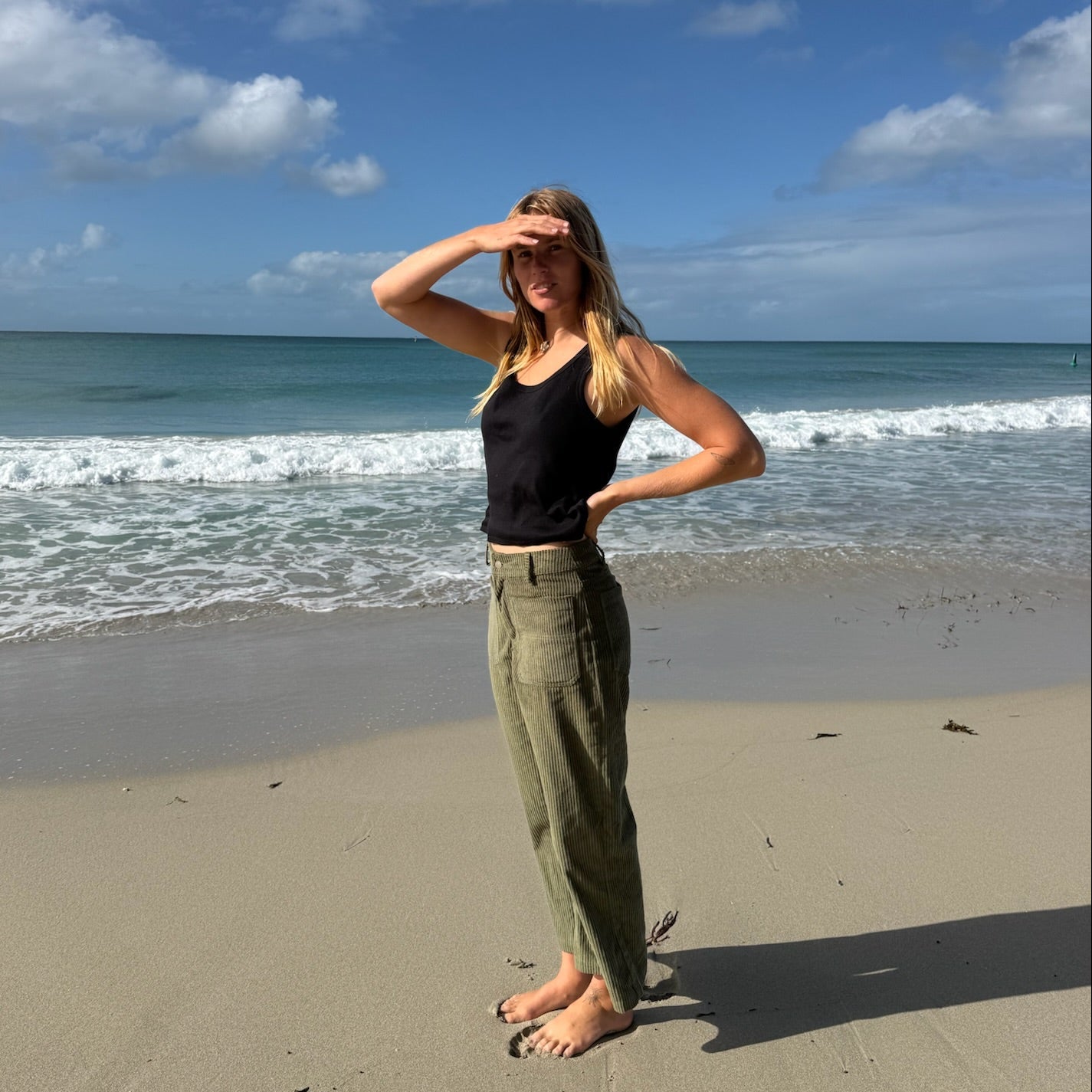 A woman stands on a sandy beach with the ocean and blue sky behind her, wearing a black tank top and olive green pants.