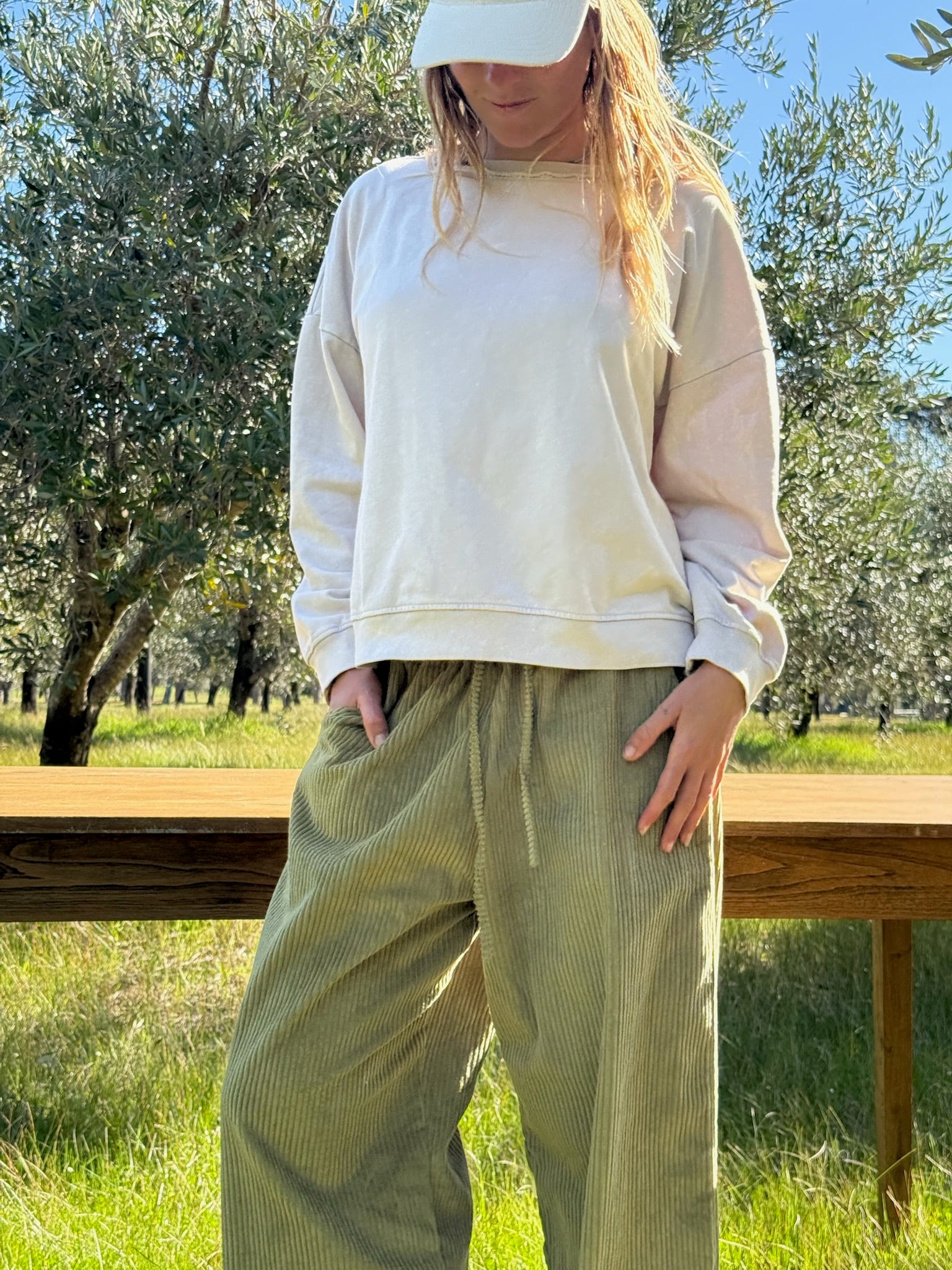 woman in cream coloured sweatshirt, cap and green corduroy pants in olive grove