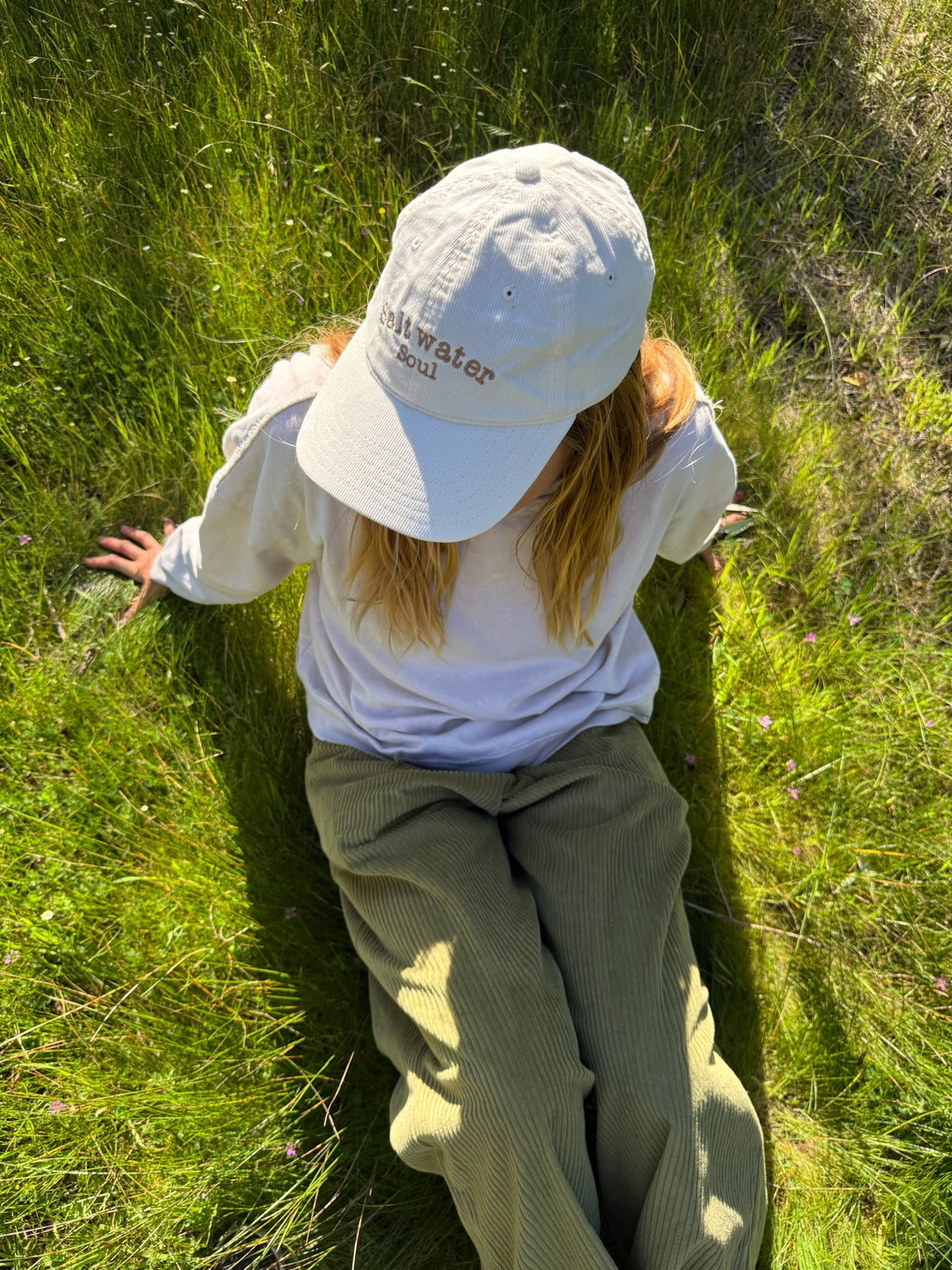 A woman wearing a white baseball cap with 'Saltwater Soul' embroidered on the front, a white sweatshirt shirt, and olive green corduroy pants, sitting on a sunny green grassy field.