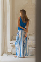 Load image into Gallery viewer, Woman in a blue top and striped pants standing in a room with white walls and furniture.
