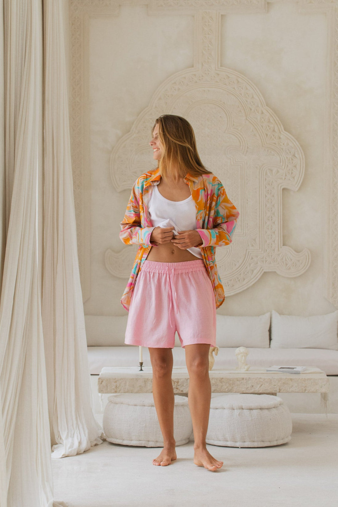 Woman in colorful shirt and pink shorts standing in a stylish room with white decor.