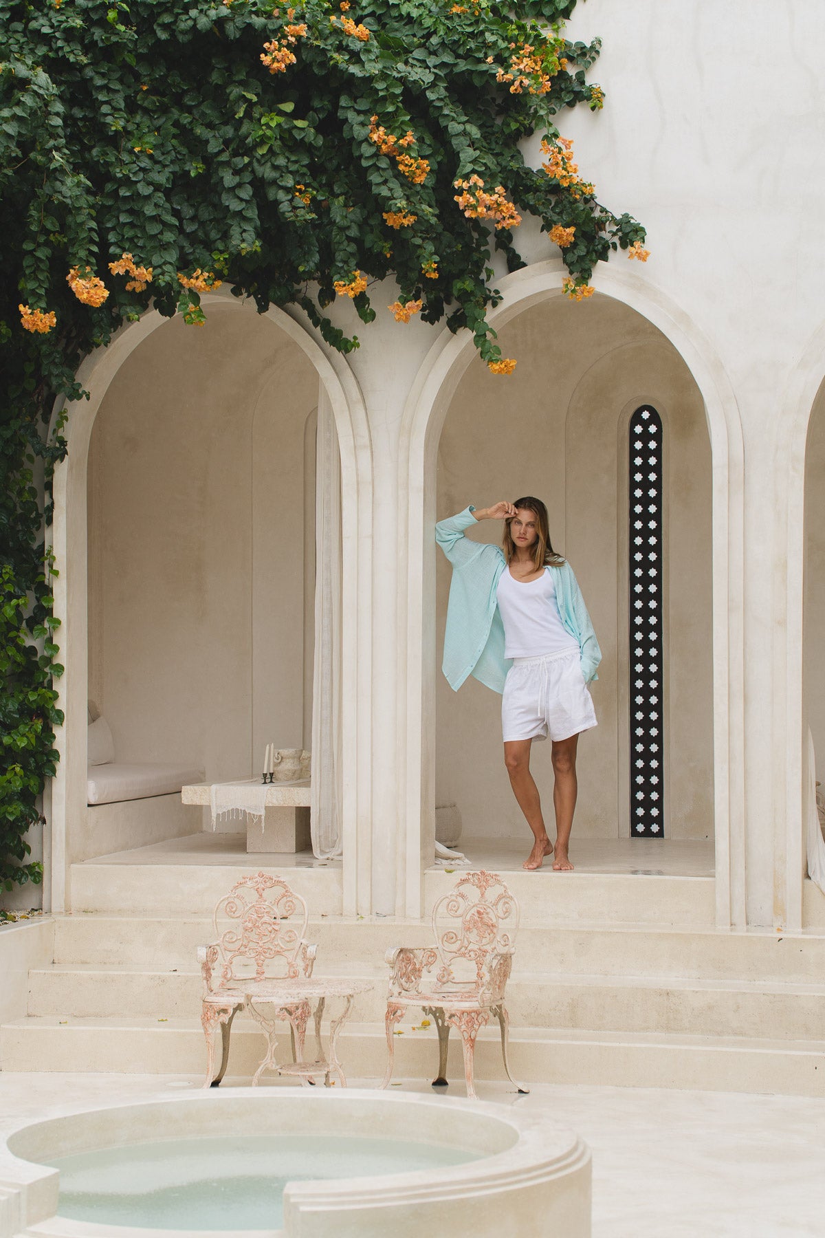Woman in a light blue shirt , white tank top and white shorts standing in an elegant outdoor setting with arches and greenery.