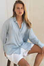 Load image into Gallery viewer, Woman wearing a light blue shirt over  a white tank top and white shorts sitting on a stool against a neutral wall.
