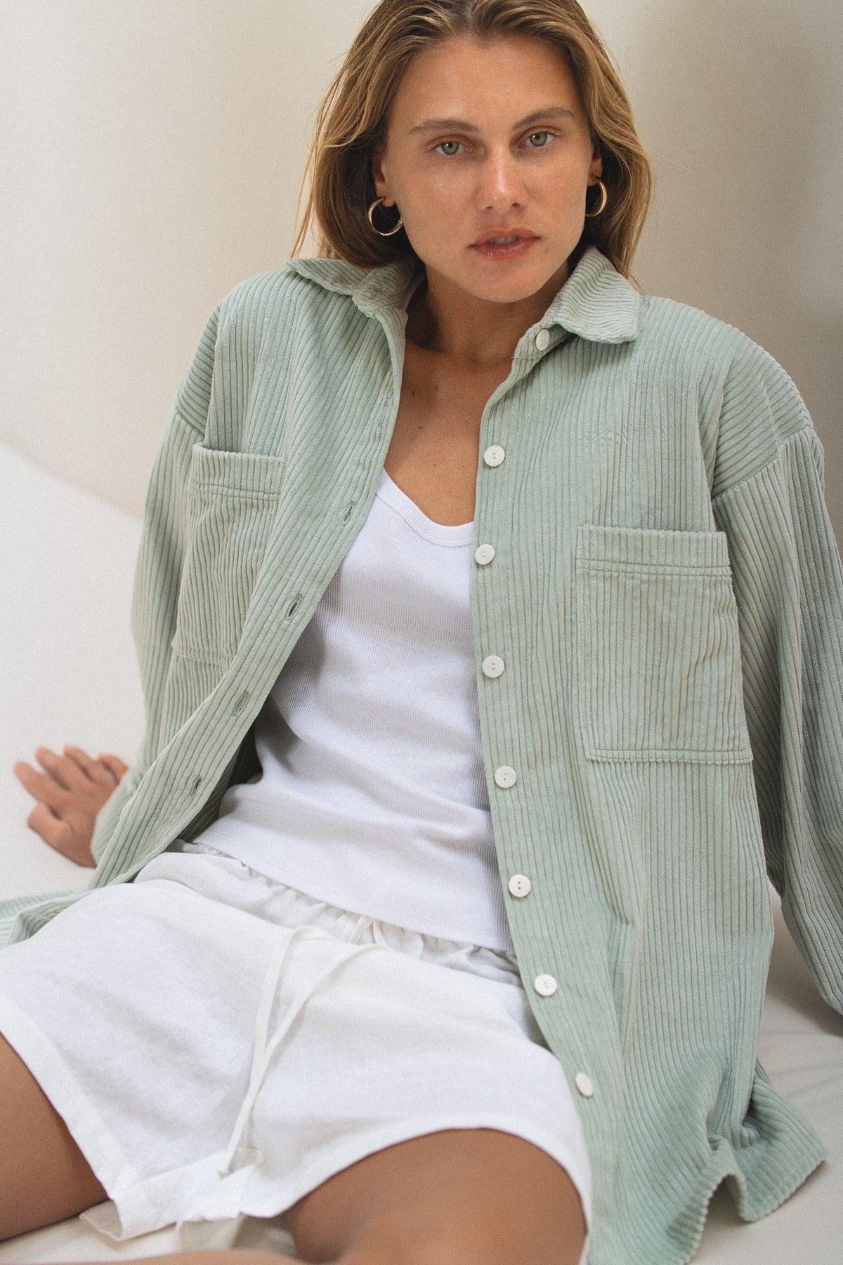 Woman wearing a light green corduroy shirt over a white top and shorts, sitting on a couch.