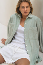 Load image into Gallery viewer, Woman wearing a light green corduroy shirt over a white top and shorts, sitting on a couch.
