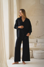Load image into Gallery viewer, Woman in a black shirt, tank top and pants standing in a luxurious room with white decor
