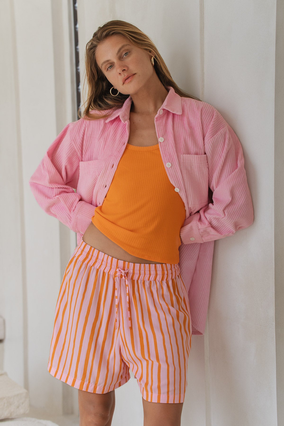 Woman wearing a pink jacket, orange top, and orange striped shorts against a white wall.