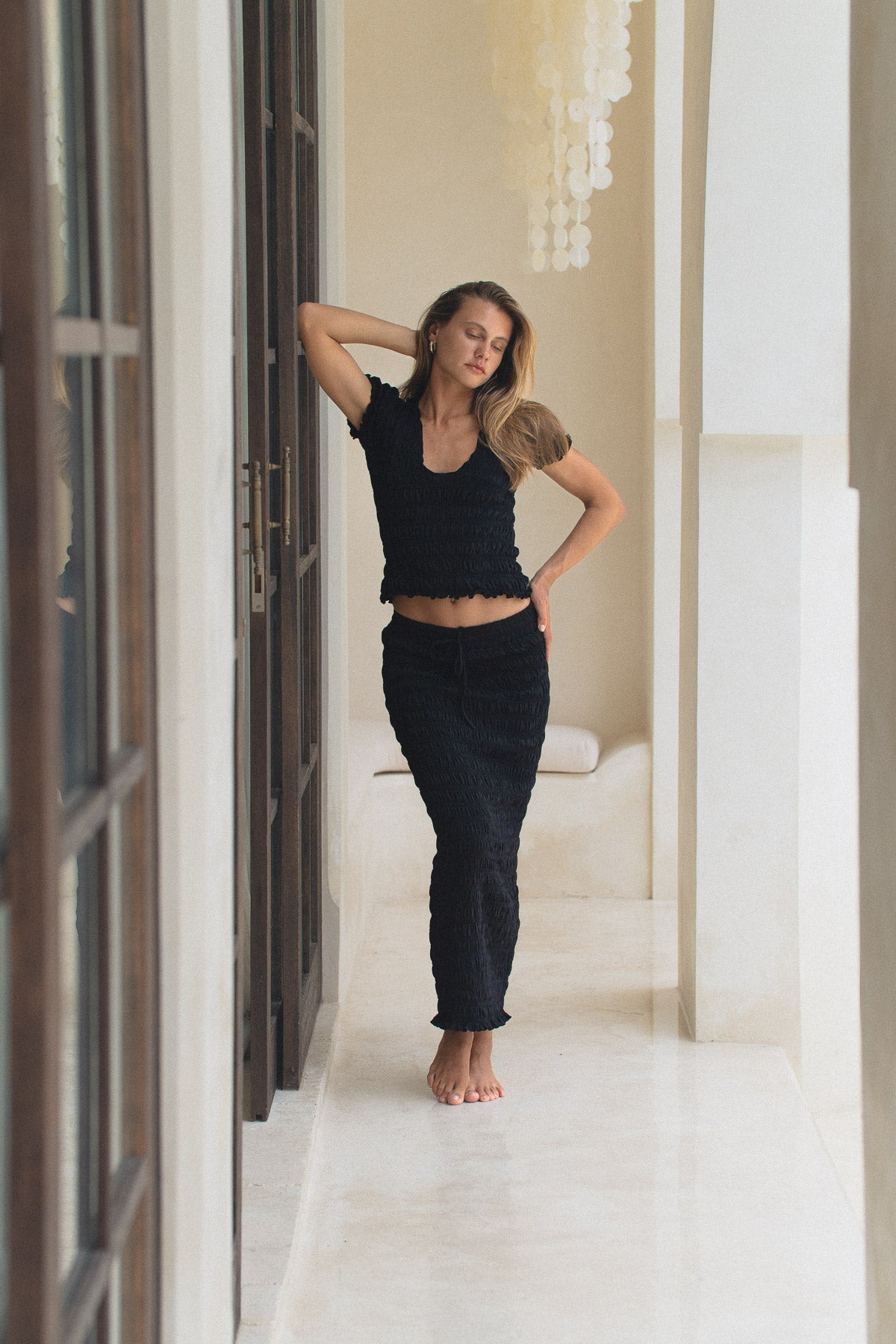 Woman in black shirred top and shirred maxi skirt standing in a bright room with white walls and a chandelier.