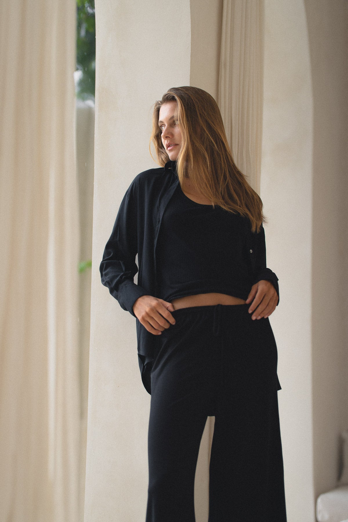 Woman wearing a black shirt, black tank and pants standing in a softly lit room with curtains.