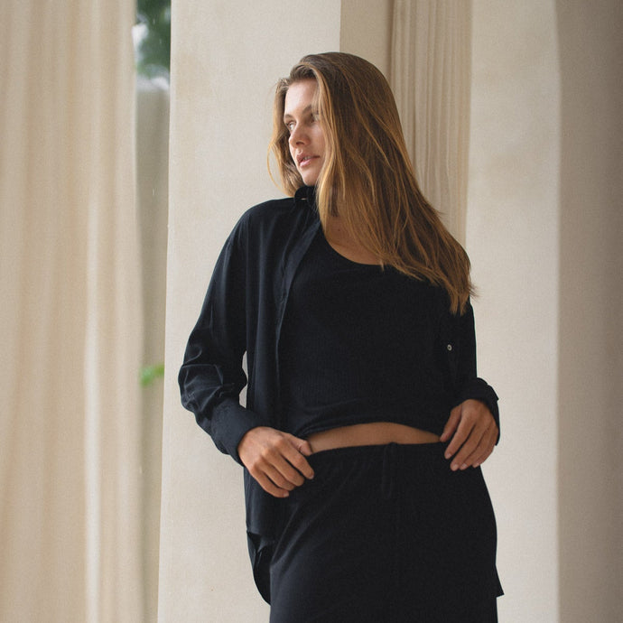 Woman wearing a black shirt, black tank and pants standing in a softly lit room with curtains.