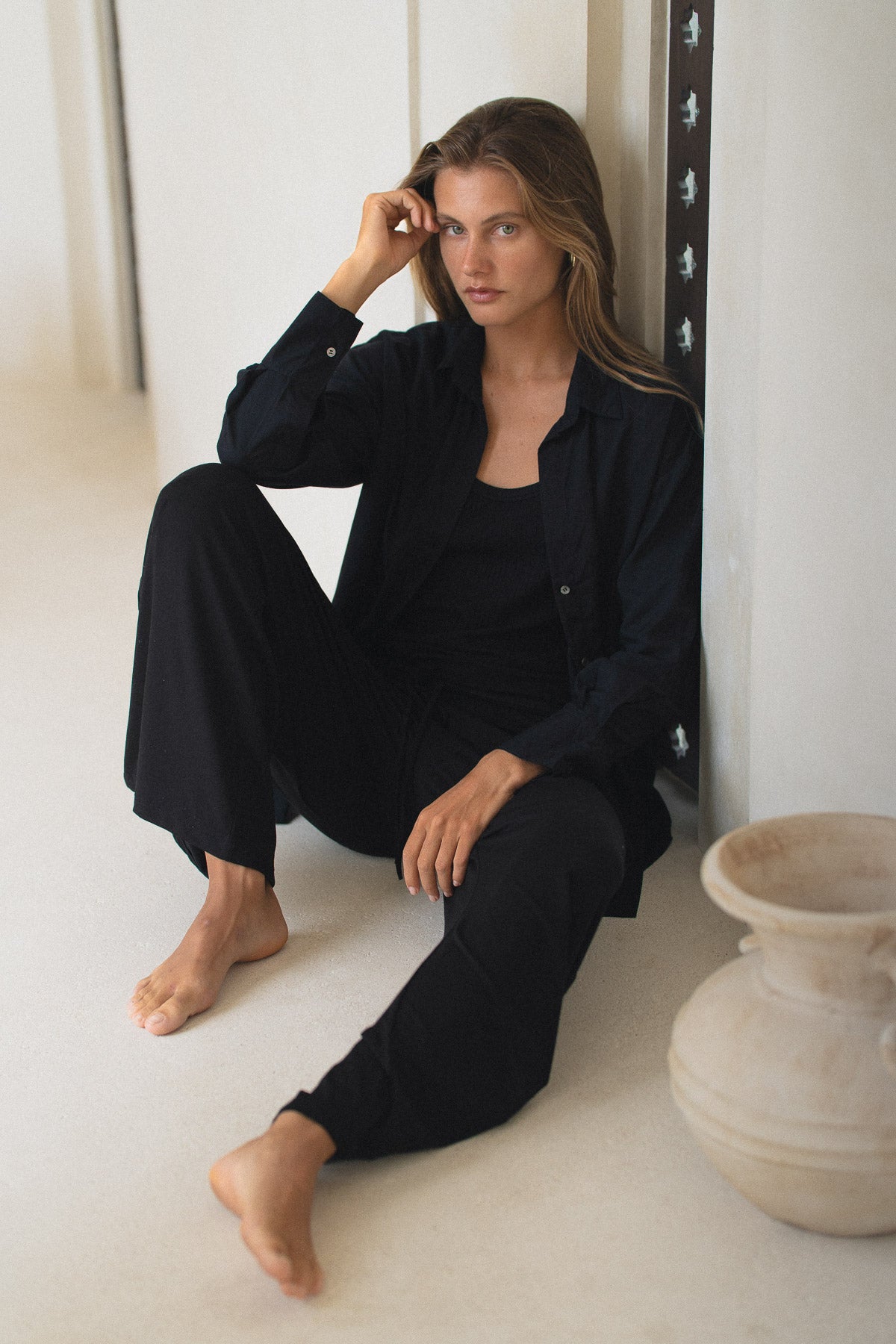 Woman wearing a black outfit sitting on a white floor with a vase in the background