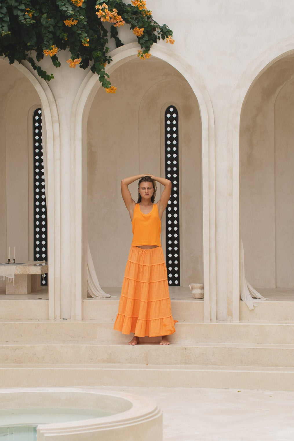 Woman in an orange skirt and tank top standing in front of a classical architectural archway in a marble courtyard.
