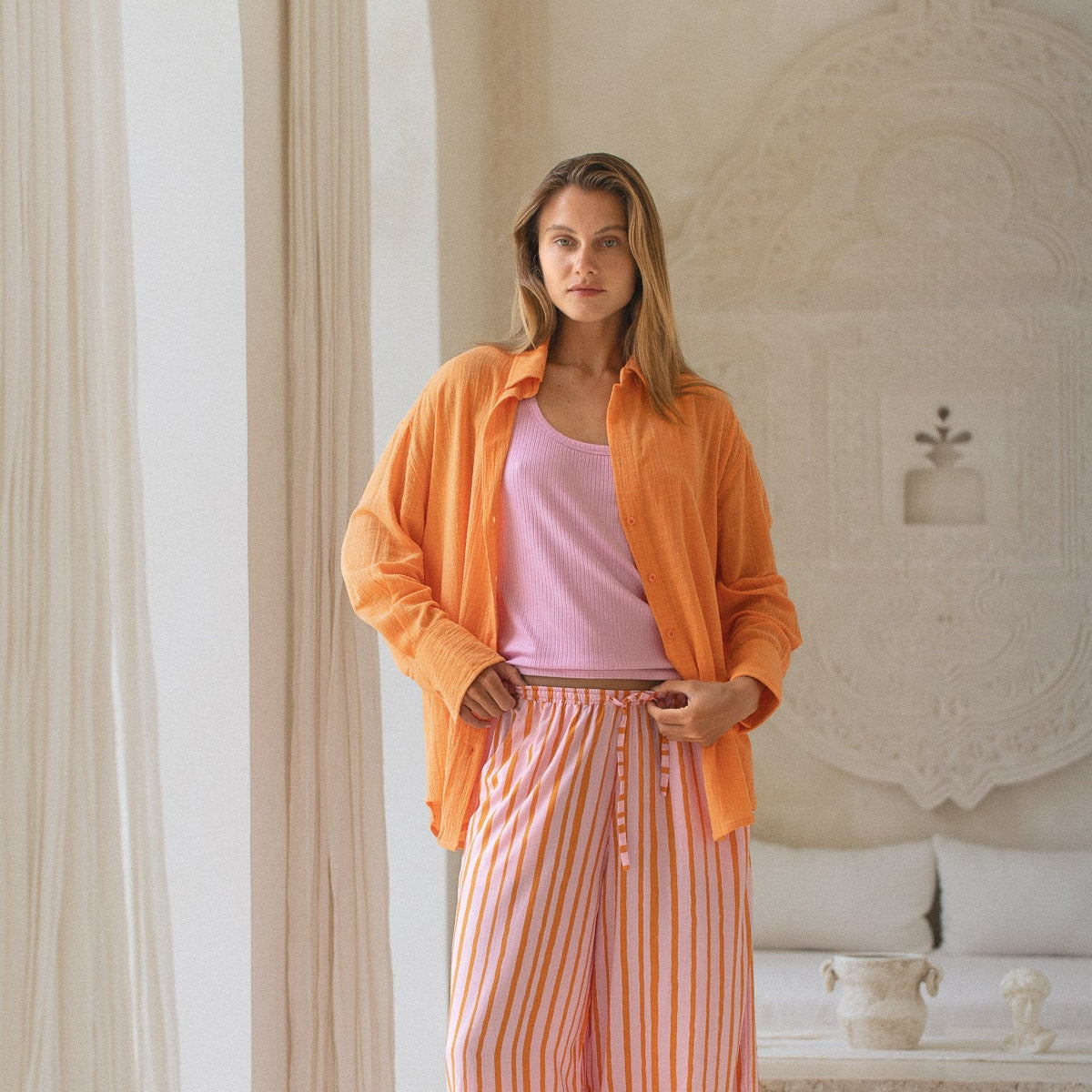 Woman wearing an orange shirt and pink striped pants in a bright room.