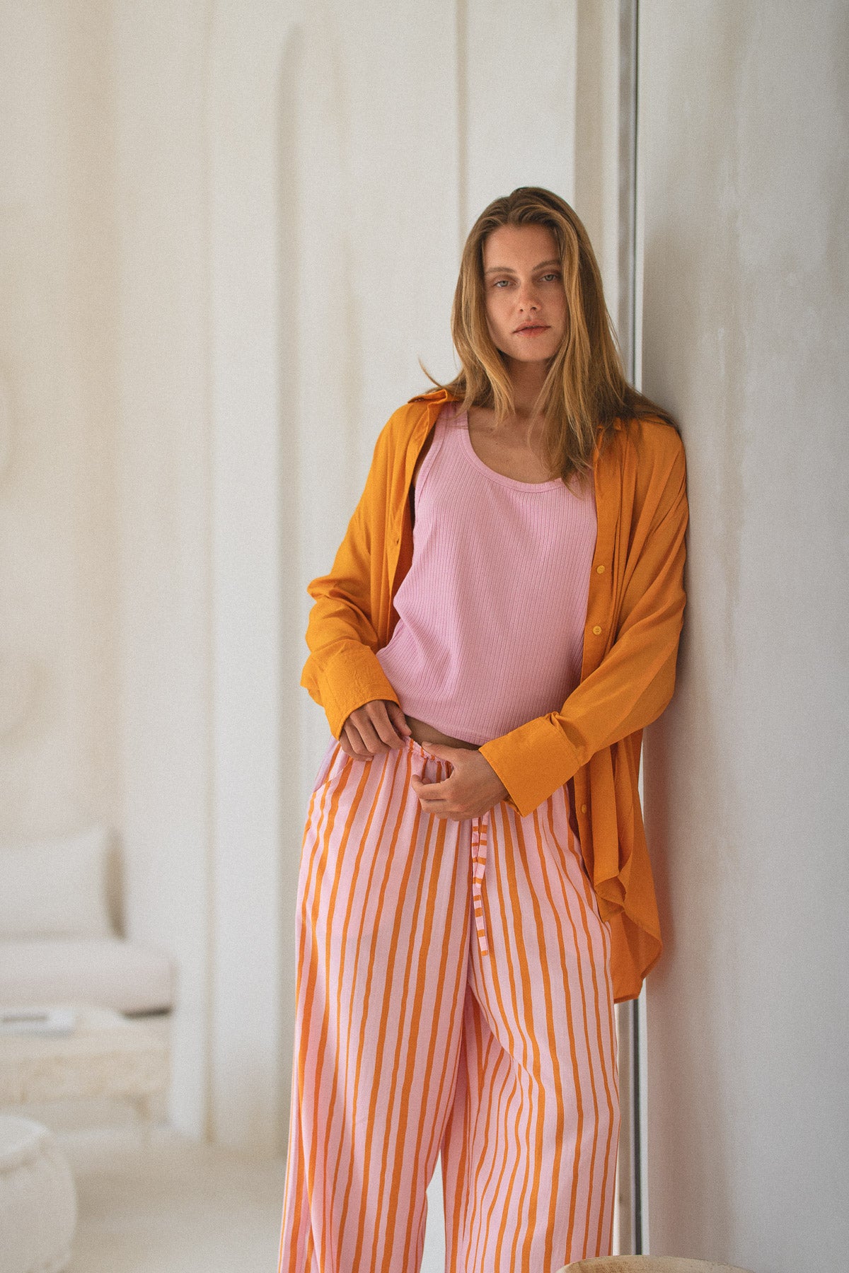 Woman wearing a pink tank top and orange striped pants leaning against a white wall.