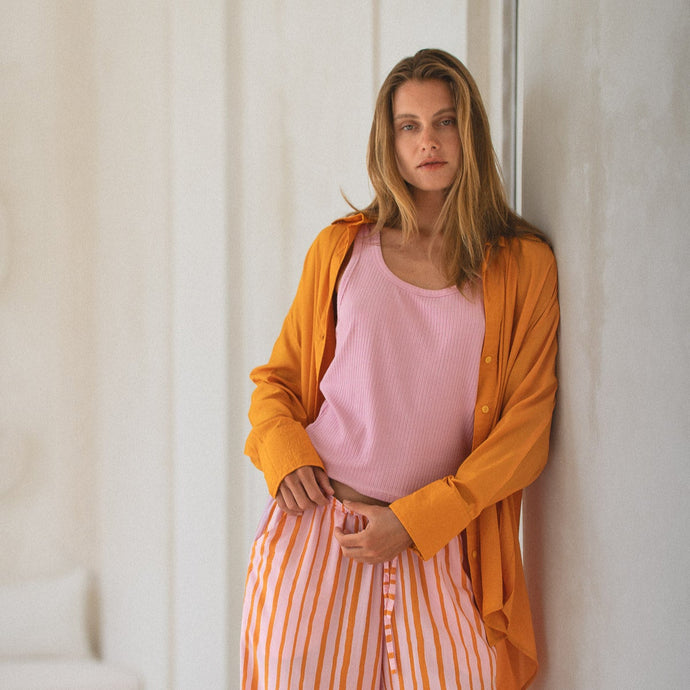 Woman wearing an orange shirt over a pink top and orange striped pants leaning against a white wall.