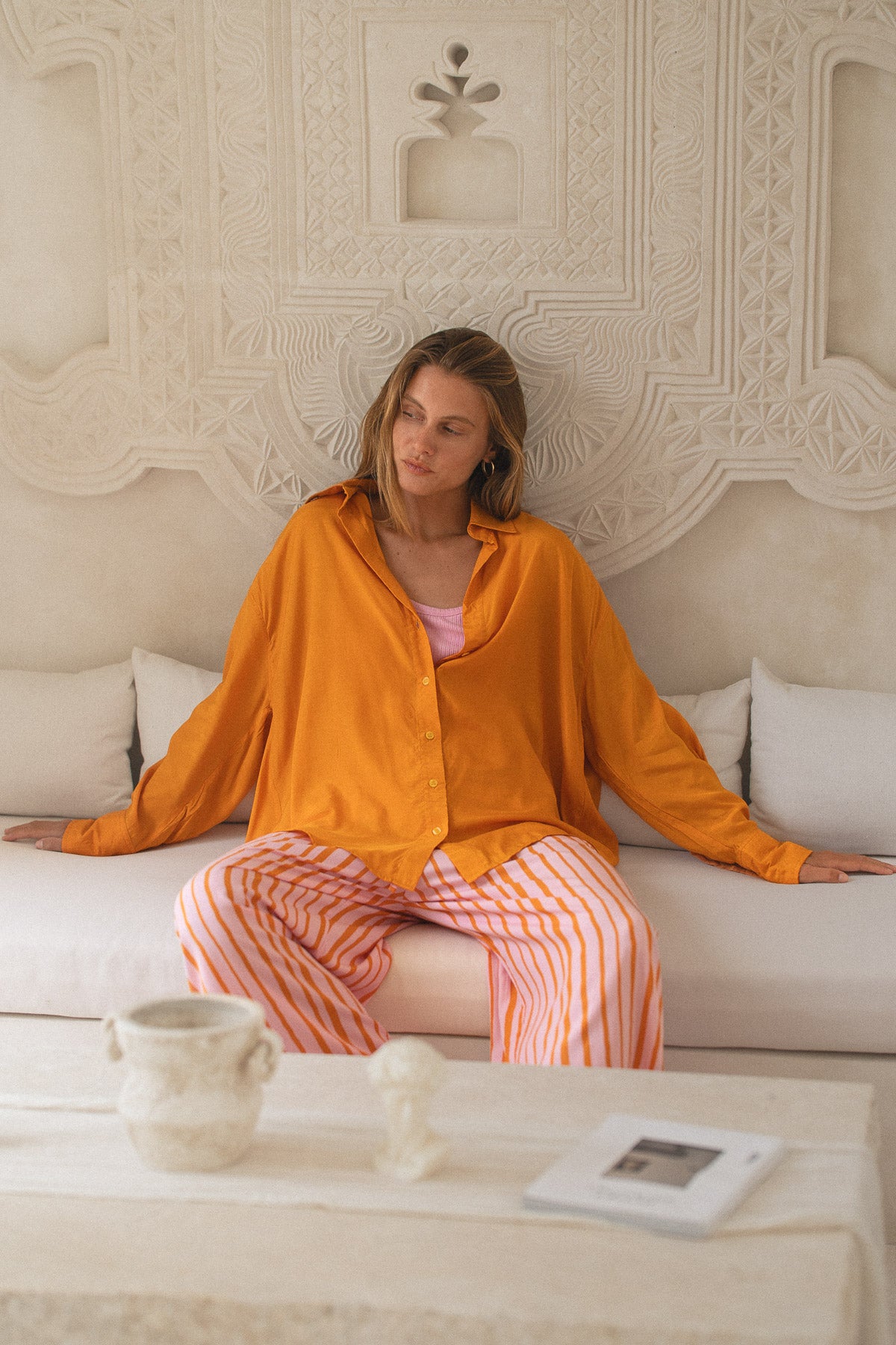 Woman in orange shirt and pink pants sitting on a couch in a decorated room.