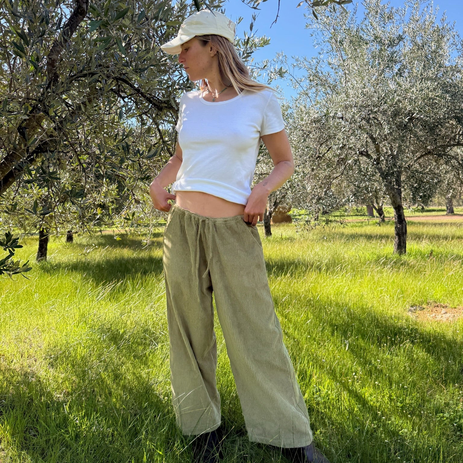 A woman stands in a grassy outdoor olive grove wearing a white crop top, olive green wide-leg pants, a beige baseball cap, and dark boots.