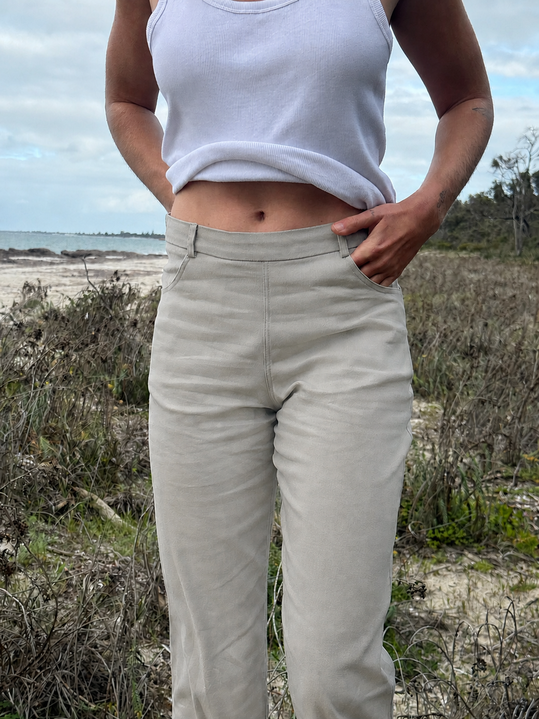 A person standing outdoors near a beach and shrubs, wearing light beige straight-leg pants and a white ribbed tank top.
