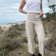 A person standing outdoors on a beach path wearing light beige cropped wide-leg pants, a white tank top, black socks, and black shoes.