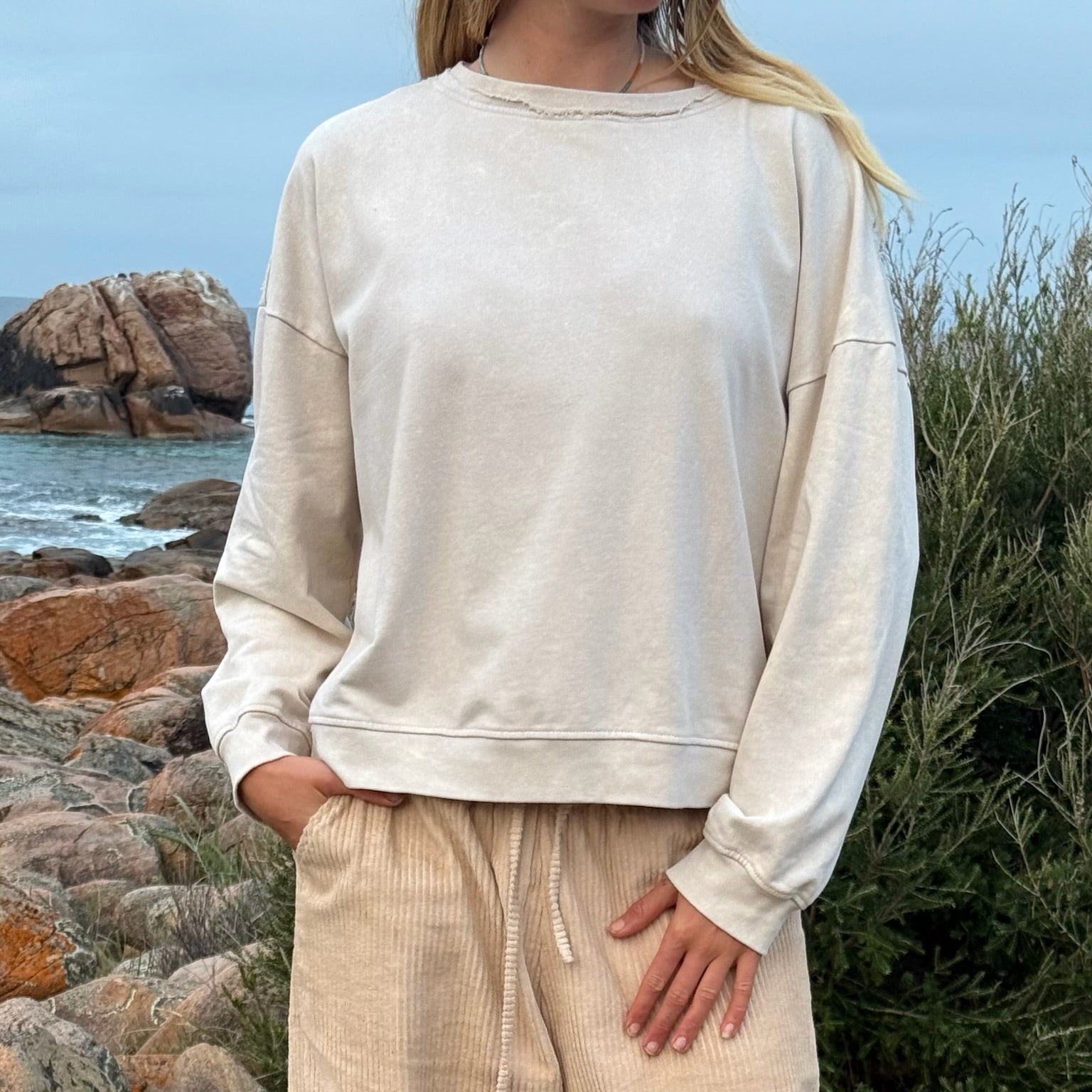 A woman wearing a cream-colored crewneck sweatshirt and matching corduroy pants, standing outdoors on a rocky beach with the ocean and sky in the background.