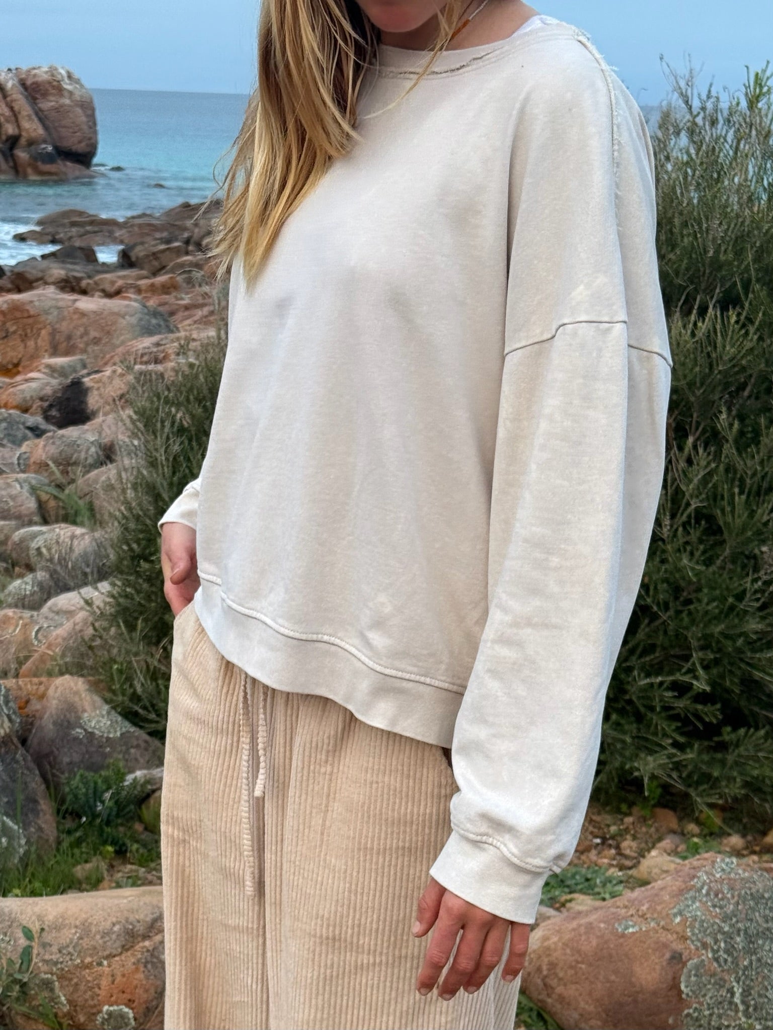A woman wearing an oversized cream-colored sweatshirt and beige corduroy pants, standing outdoors on a rocky beach with the ocean and greenery in the background.