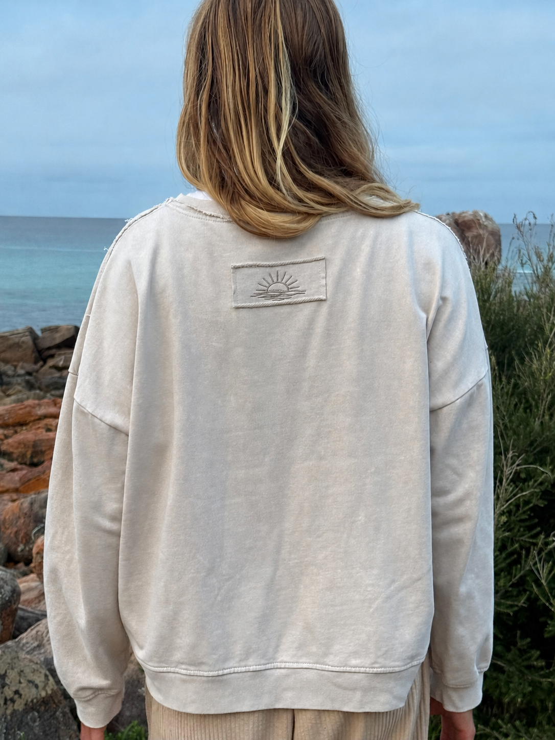A person viewed from behind wearing a light beige crewneck sweatshirt with a small embroidered sun patch on the back, standing outdoors near the ocean.