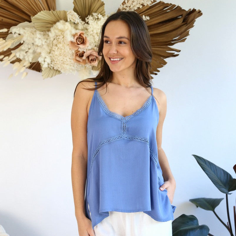 Woman wearing a blue tank top and white pants standing in front of decorative plants and flowers.