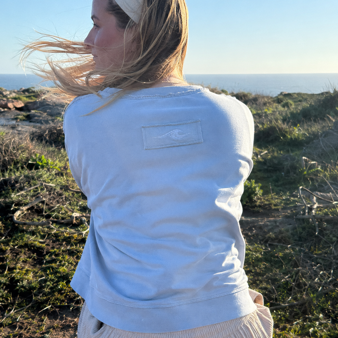 A woman viewed from behind wearing a light blue long-sleeve top and beige pants, standing outdoors on a grassy hill with the ocean and blue sky in the background.