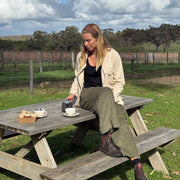 A woman sits at a rustic wooden picnic table outdoors in a grassy field under a cloudy sky, pouring tea from a metal pot. She is wearing a beige jacket, black top, olive green pants, and brown boots.