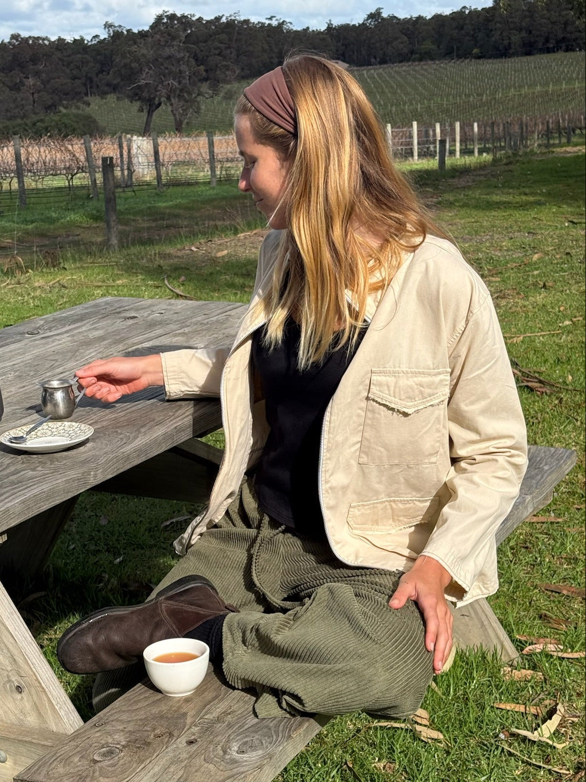A woman sits on a wooden picnic table outdoors in a grassy field with vineyard rows in the background. She is wearing a beige overshirt over a black top and olive green pants, with coffee cups on the table.