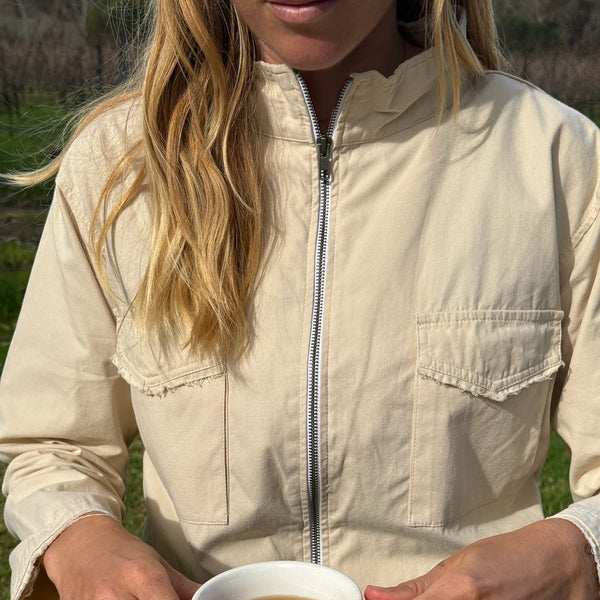 A woman wearing a light beige zip-up utility jacket sits at a wooden table outdoors, holding a white cup of tea on a patterned saucer.