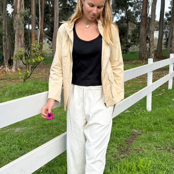 A woman standing outdoors in a grassy field next to a white wooden fence, wearing a beige jacket over a black tank top and cream-colored linen pants.