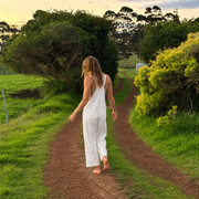 A woman in a white sleeveless jumpsuit walks barefoot down a dirt path surrounded by green grass and trees at sunset.
