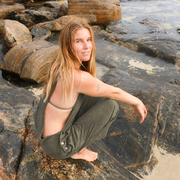 A woman crouching on dark rocks by the water, wearing an olive green overalls.