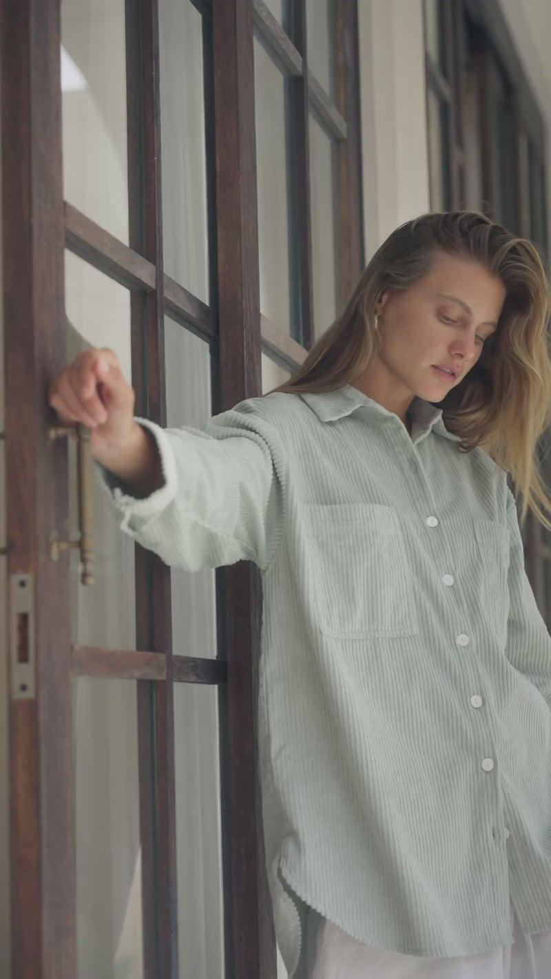 Woman standing in a passageway wearing a light green corduroy shirt and white shorts.