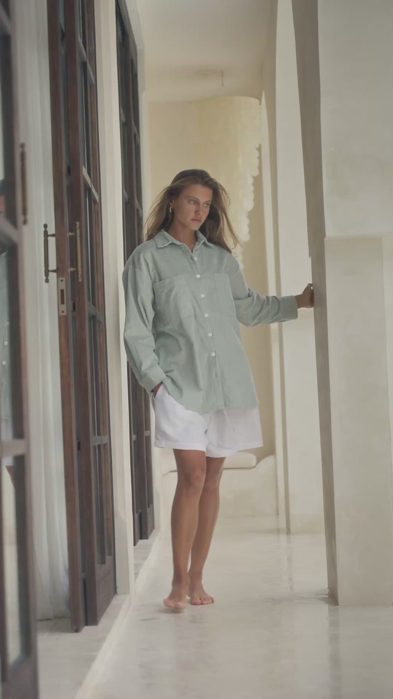 Woman standing in a passageway wearing a light green corduroy shirt and white shorts.