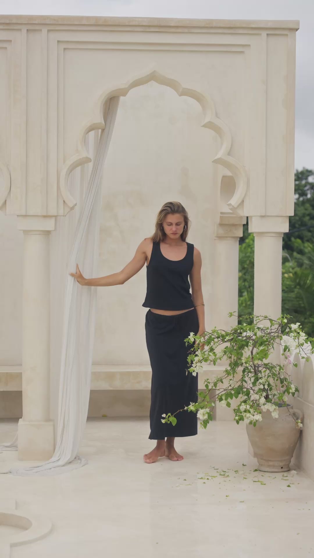 Load video: Woman in a black tank top and skirt  standing on steps with white columns in the background