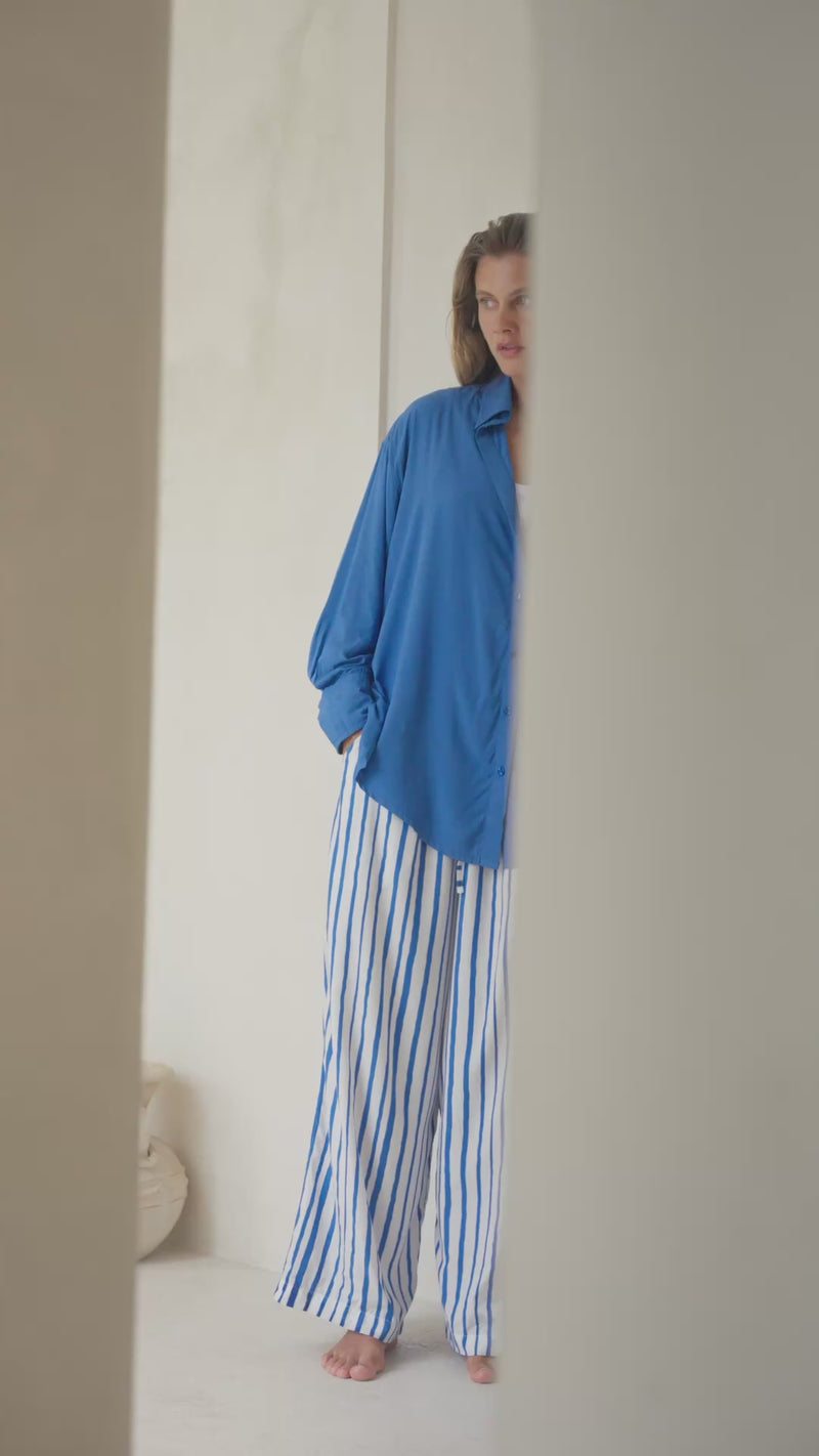 Woman standing by a  white wall wearing a blue shirt, white tank top, and striped pants.