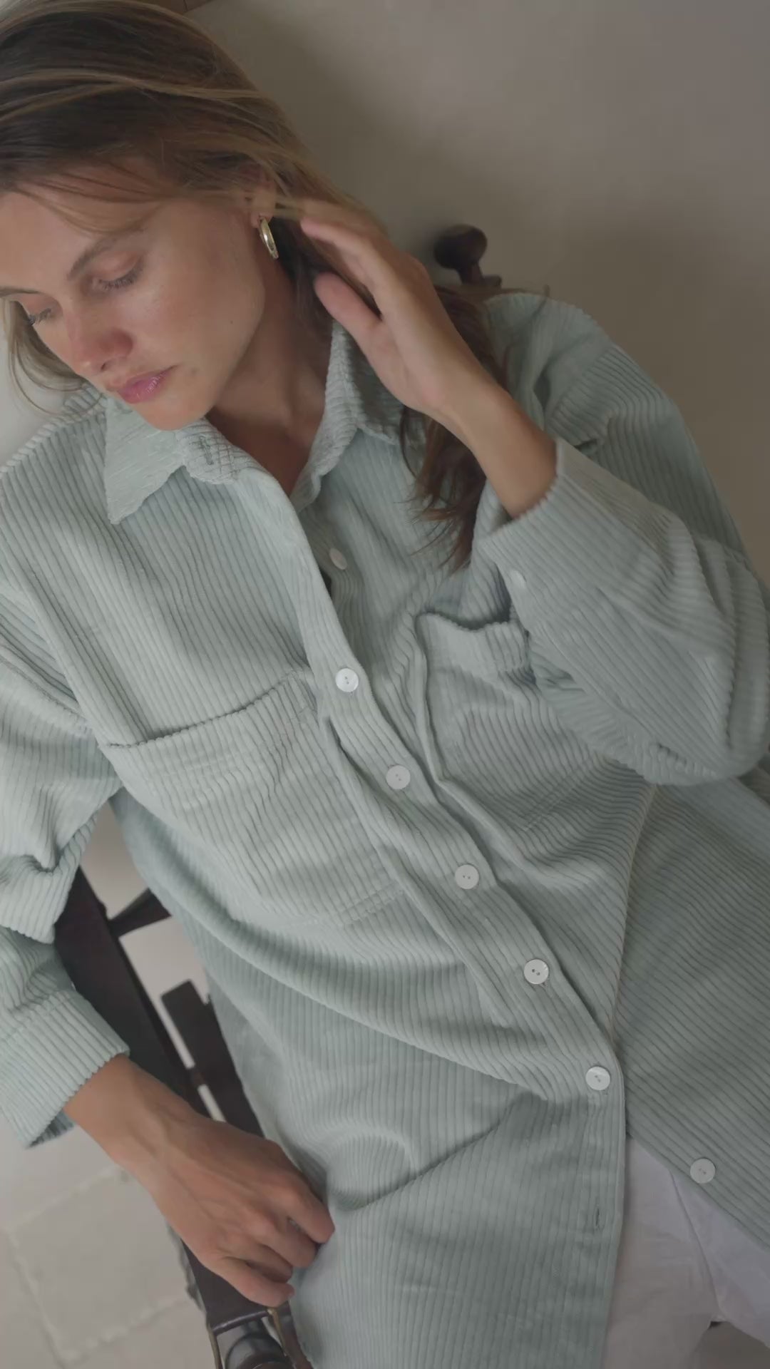 Load video: Woman sitting in a softly lit room wearing a light green corduroy jacket and white shorts.